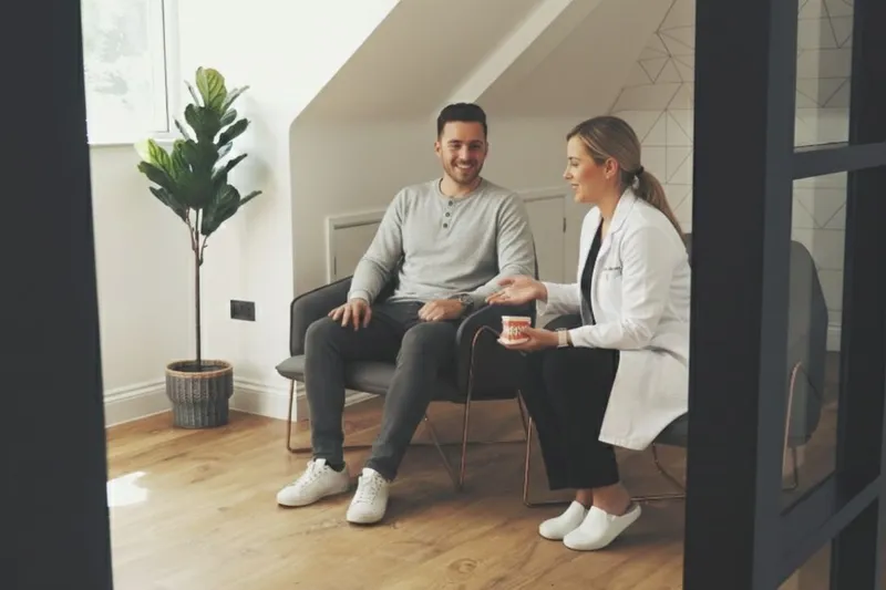 A female dentist in a white clinical coat sitting in a modern, calm waiting area, leaning forward to explain a dental model to a smiling male patient.