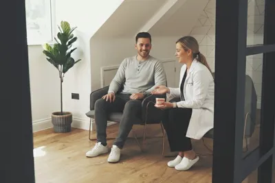A female dentist in a white clinical coat sitting in a modern, calm waiting area, leaning forward to explain a dental model to a smiling male patient.