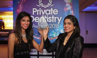 Dr. Shabnam Zai and Dr. Nikita Mehta smiling and holding up a glass trophy in front of a blue 'Private Dentistry Awards 2018' backdrop at an evening awards ceremony.