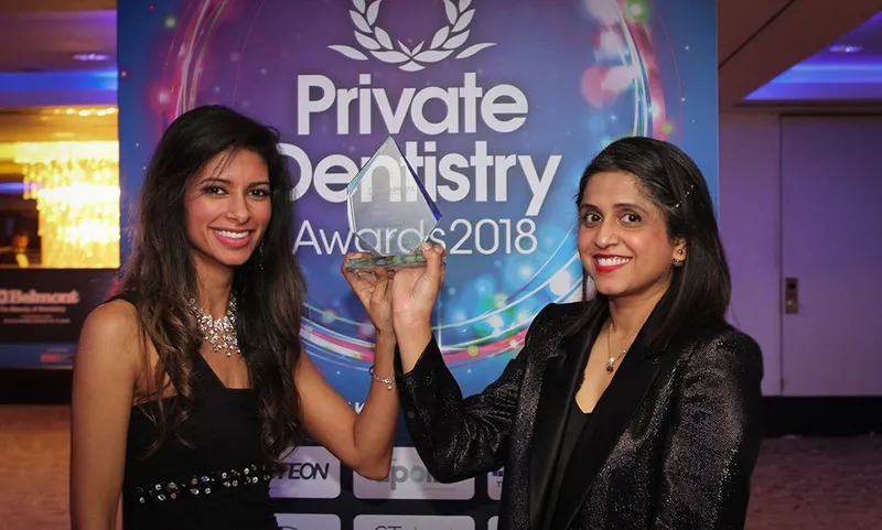 Dr. Shabnam Zai and Dr. Nikita Mehta smiling and holding up a glass trophy in front of a blue 'Private Dentistry Awards 2018' backdrop at an evening awards ceremony.